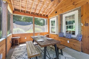 A wooden interior with a table set for dining, a bench with cushions, and large windows offering a view of greenery.