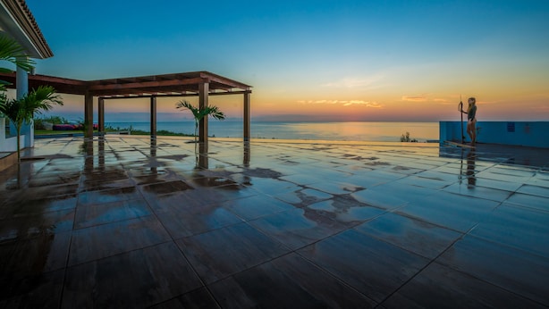 Un amplio espacio al aire libre con una pérgola de madera, suelo de baldosas y vistas al mar al atardecer.