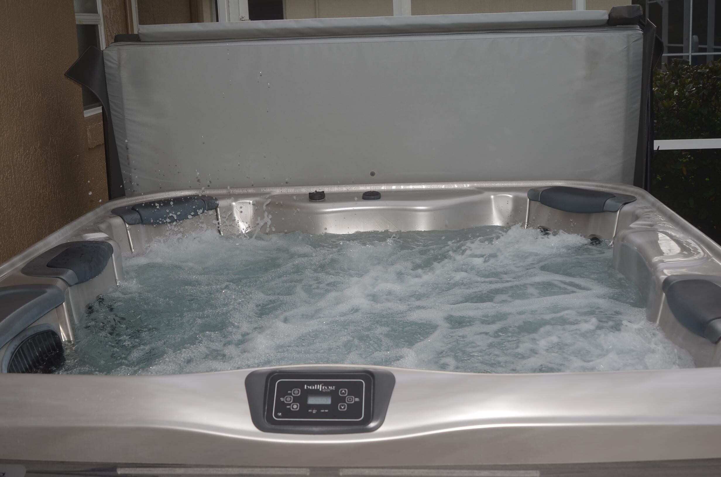 A hot tub with a control panel, surrounded by a white exterior wall and a window.