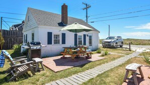 Une maison blanche avec une cheminée en briques, un barbecue et une table de pique-nique avec un parasol. Sur la terrasse, on trouve des chaises en bois et une petite table.