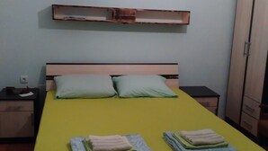 A bed with a yellow bedspread, two pillows, and folded towels on the bed. There is a wooden nightstand with a lamp, a wooden wardrobe, and a shelf above the bed.