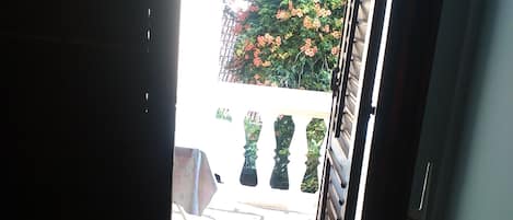 Uma porta aberta dá para um pátio com piso de pedra e vista de um jardim florido com cerca.