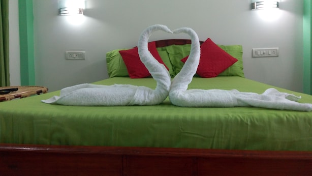 A bed with a green bedspread and red pillows, two white towels arranged in a heart shape, and two wall-mounted lights.