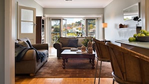 A cozy living room with a sofa, a coffee table, and a dining area with chairs. There is a large window providing a view of the outside. - Jesmondene Daylesford (Daylesford)