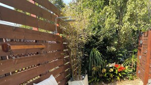 A wooden deck with a wicker sofa and a glass table, surrounded by lush greenery and a wooden fence.