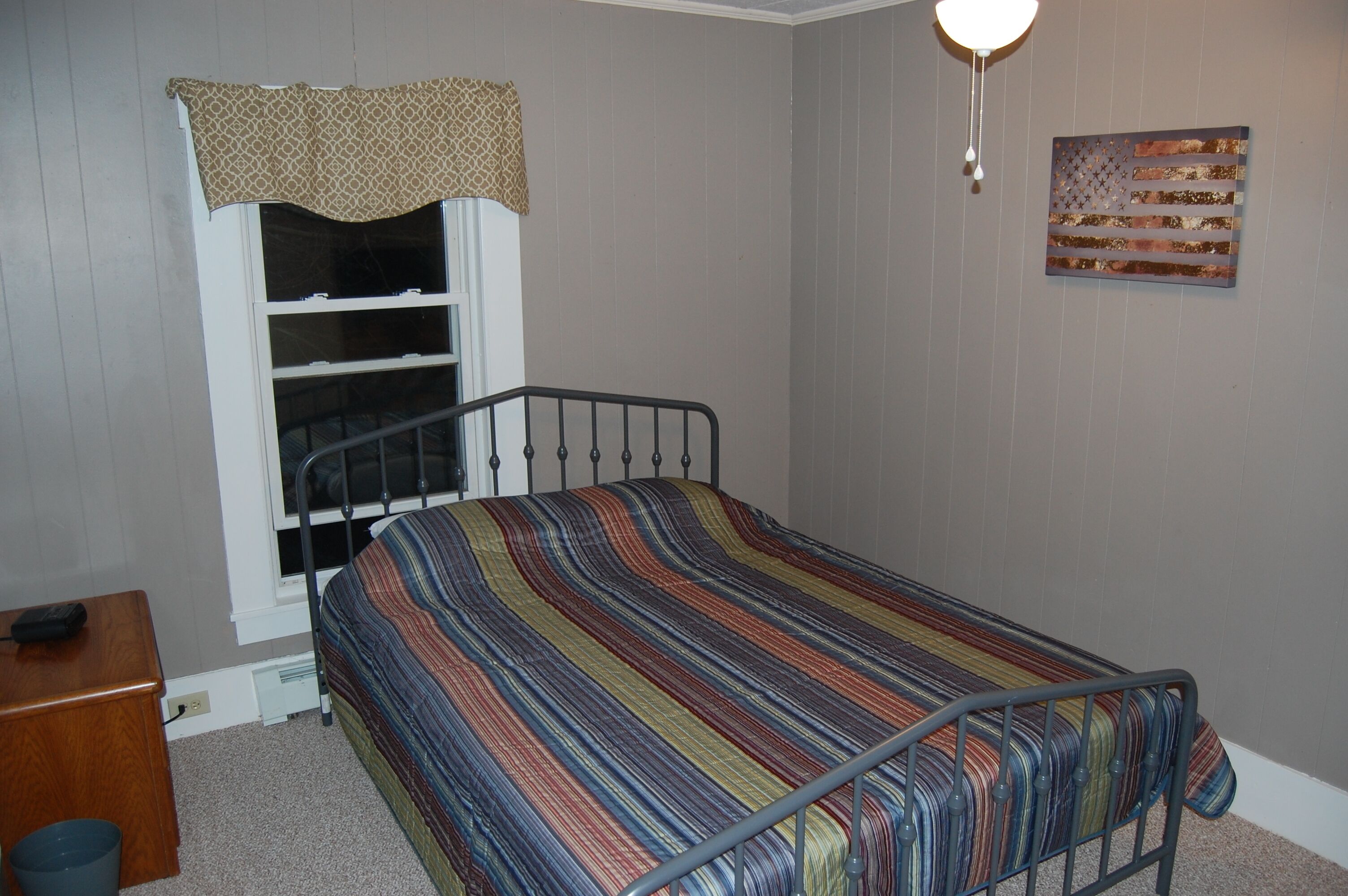 A small, single-bed room with a metal frame, a window with a patterned valance, a wall-mounted American flag, and a bedside table with a lamp.