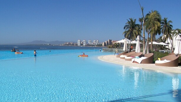 Una amplia piscina con agua cristalina y azul, rodeada de sillas de descanso y sombrillas, con vistas al océano y edificios a lo lejos.