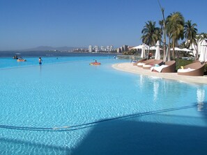 A large swimming pool with clear blue water, surrounded by lounge chairs and umbrellas, and a view of the ocean and distant buildings. -  PENINSULA-SPECTACULAR OCEAN AND MOUNTAIN VIEWS, HUGE BEACHFRONT INFINITY POOL (PUERTO VALLARTA)