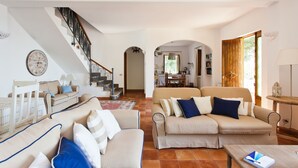A spacious living area with a sofa set, a staircase, and an open kitchen in the background.