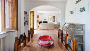 A spacious living room with a wooden floor, a central glass table, and a sofa. There are bookshelves and a television in the background.