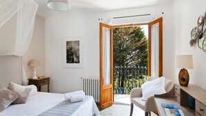 A bedroom with a bed, a chair, a desk, a radiator, and a balcony with a view of trees.