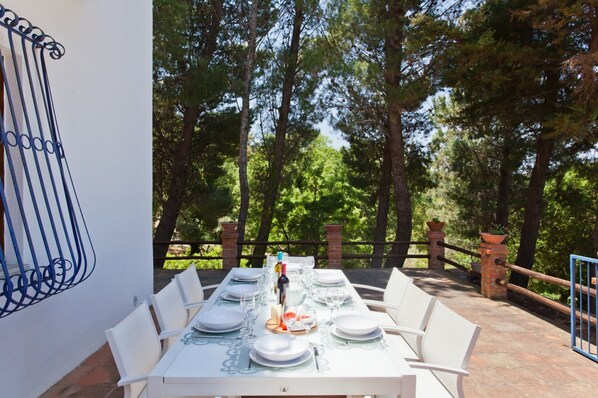 An outdoor dining area with a table set for four, surrounded by white chairs, with a view of tall trees in the background.