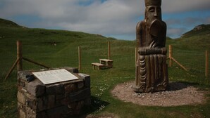 A stone statue of a figure with a crown, set in a grassy field with a stone bench and a sign in the foreground. - An Airigh  - within walking distance from Stornoway (Stornoway, Isle of Lewis, Scotland)