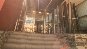 A modern building with a glass facade, a set of stairs leading to a covered entrance, and a tall structure in the background. - Hotel Estilo MB (Villa Carlos Paz)