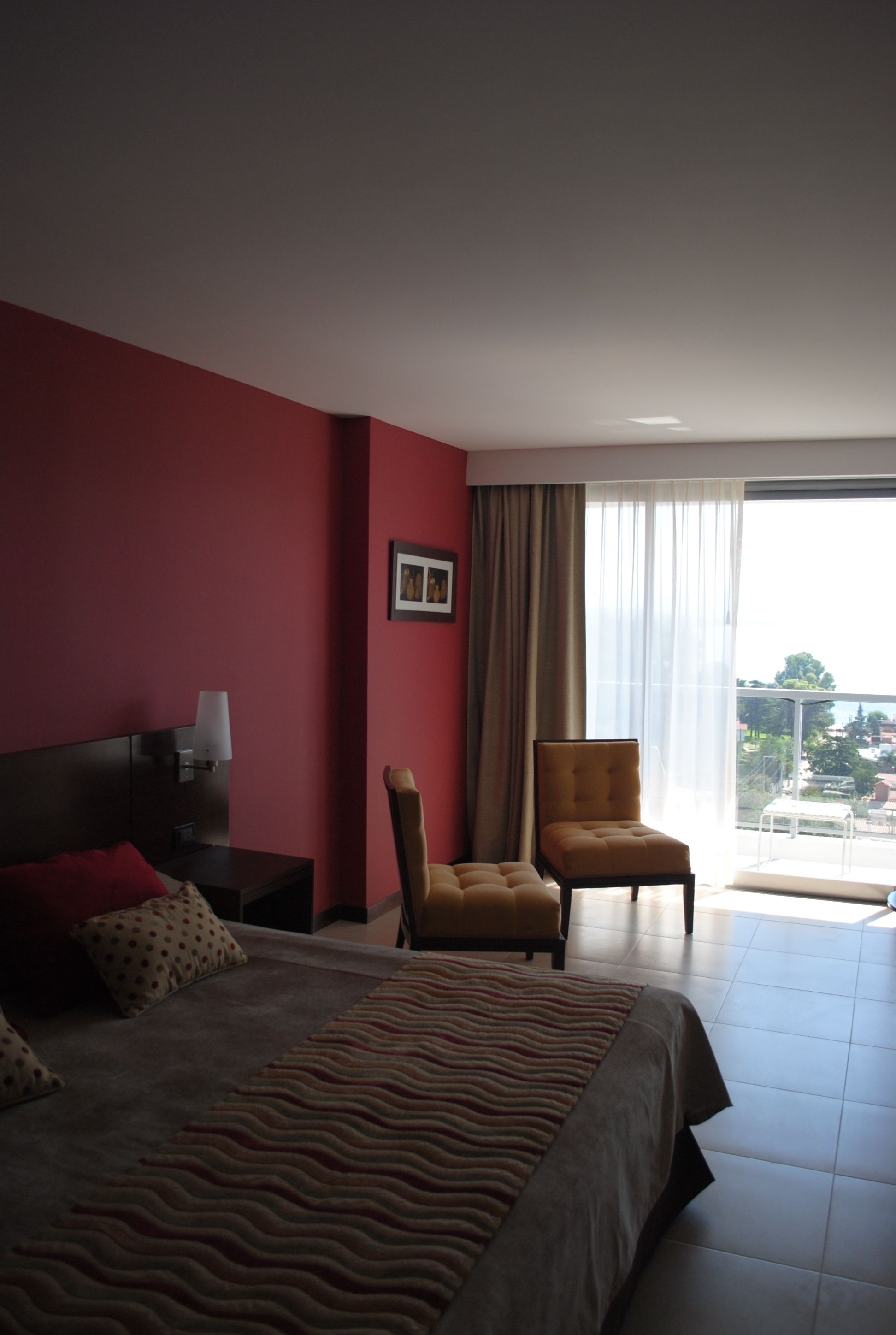 Habitación de hotel con cama, silla, mesita de noche, lámpara y ventana con vistas.