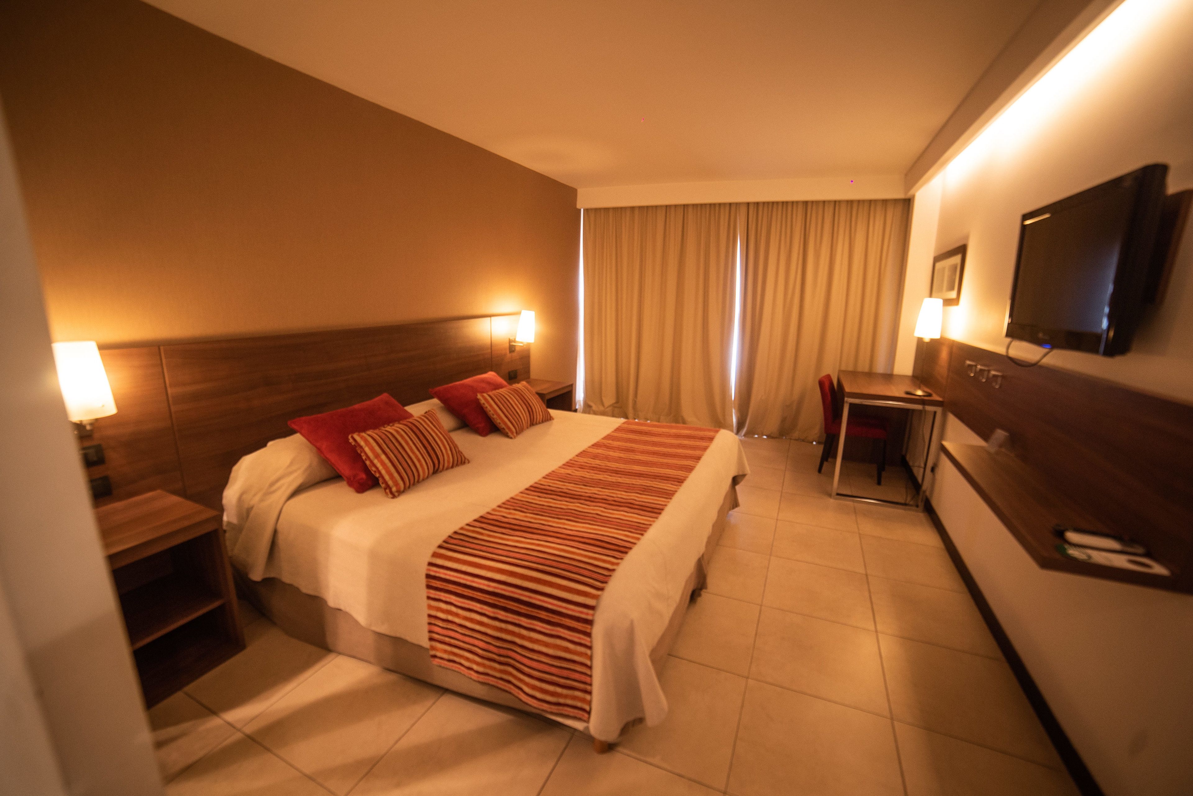 Habitación de hotel con cama, mesitas de noche, escritorio, silla, televisión y ventana con cortinas.
