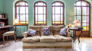 Une pièce chaleureuse avec un canapé beige, une chaise en bois, une petite table et trois fenêtres en arc de cercle donnant sur l’extérieur.
