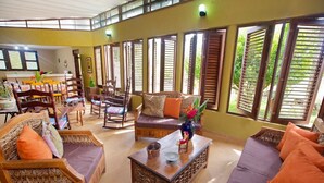 A spacious living area with a sofa, chairs, and a coffee table. There are large windows with shutters, and a dining area is visible in the background. - CASA ANACAHUITA, Limonal, Jarabacoa (Jarabacoa)