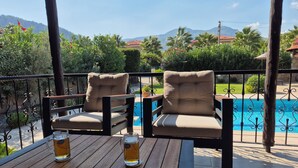Una terraza de madera con dos sillas acolchadas y dos jarras de cerveza sobre una mesa, con vistas a una piscina y exuberante vegetación.
