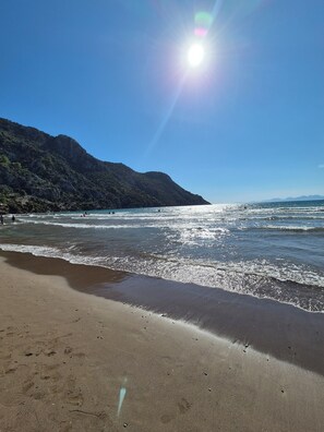A beach with clear skies and calm waters, a sandy shore, and a mountain in the background.