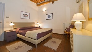 Un dormitorio con cama, lámparas de noche, un escritorio con lámpara, dos cuadros enmarcados, un radiador y techo de madera.