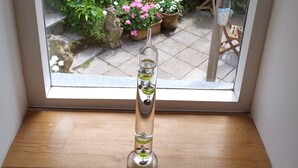 A room with a wooden floor, a window with a view of a garden, and a clear glass vase on a wooden table. - Character Cottage With Pretty, Secluded Garden (St Teath)