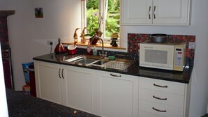 A kitchen with white cabinets, a microwave, and a window with a view of greenery. - Character Cottage With Pretty, Secluded Garden (St Teath)
