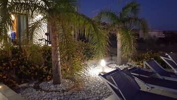 Area esterna illuminata di notte, con palme, un vialetto di ciottoli bianchi e poltrone da relax.