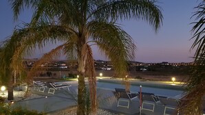 Een zwembadgedeelte met ligbedden, een palmboom en een uitzicht op een stad in de verte bij zonsondergang.