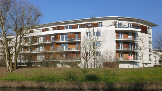 Ein mehrstöckiges Wohngebäude mit Balkonen, einer flachen Dachterrasse und einem Fluss im Vordergrund.