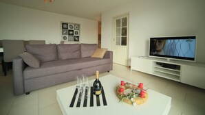Ein modernes Wohnzimmer mit einem grauen Sofa, einem weißen Couchtisch mit Gläsern und einer Flasche, einem Fernsehgerät auf einer weißen Kommode und einem dekorativen Kranz.