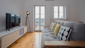 Una sala moderna con un televisor de pantalla plana, una lámpara de pie, un sofá gris con cojines estampados y un banco de madera.