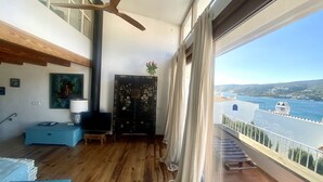 Ett rymligt rum med trägolv, en takfläkt, ett stort fönster med gardiner, utsikt över havet och en balkong med ett räcke.