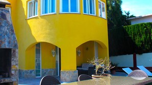 A yellow building with a stone fireplace, a covered patio with chairs, and a potted plant.