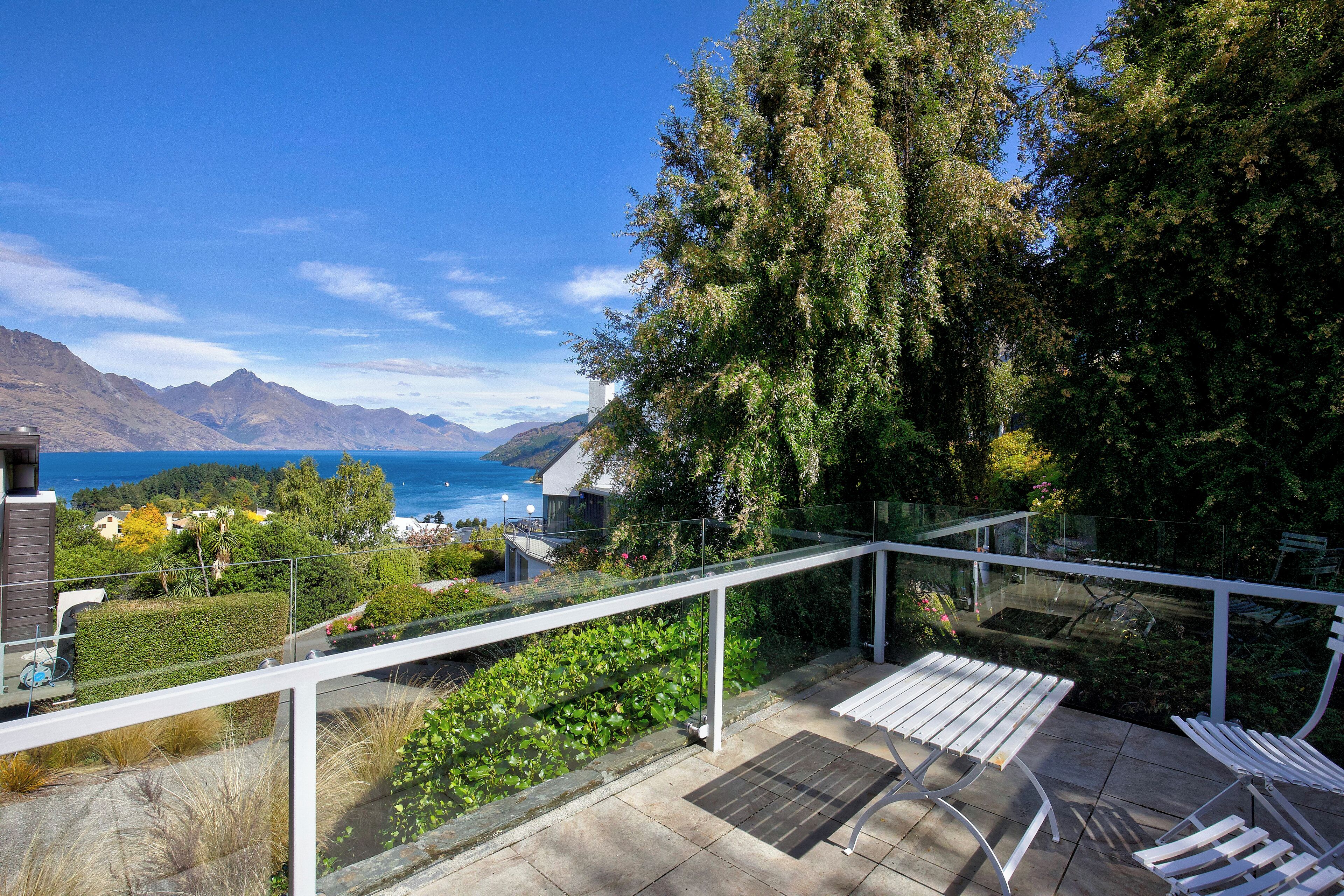 Ein Balkon mit Blick auf einen See und Berge, einer Glasreling und einer Sitzecke mit weißen Stühlen.