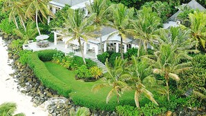 Propiedad costera con césped cuidado, palmeras y un edificio blanco con terraza cubierta.