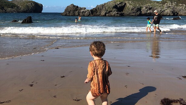 A beach scene with a child walking on the sand, waves crashing onto the shore, and rocky cliffs in the background. - Casa Dolores for 6 people (Cabrales)