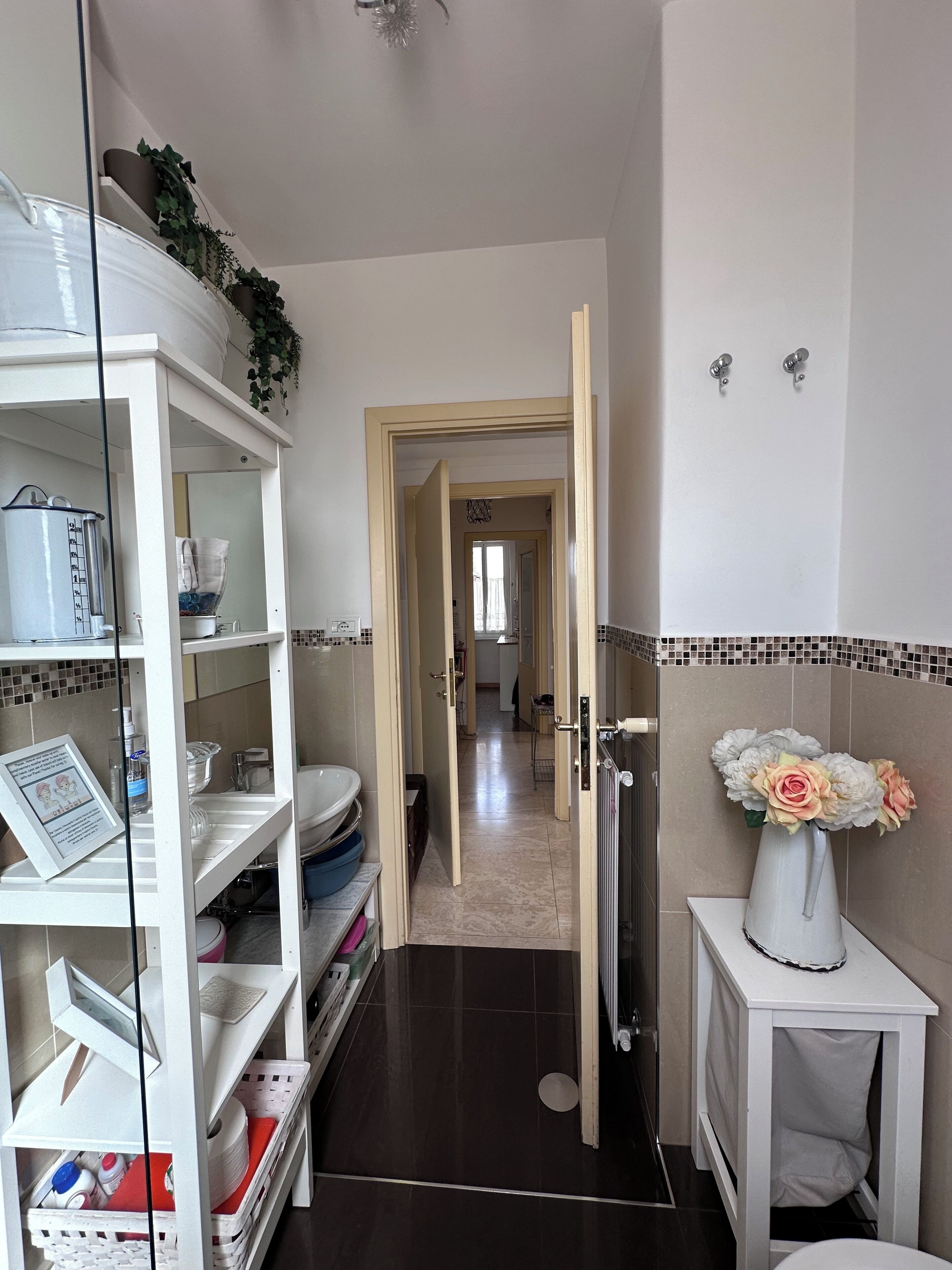 A narrow hallway with a white shelf unit on the left, a white cabinet on the right, and a door leading to another room.