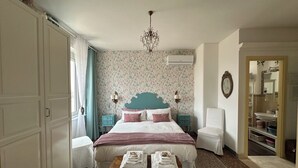 Un dormitorio con cama, mesitas de noche, una silla, una mesa de madera con toallas, una araña y un espejo.