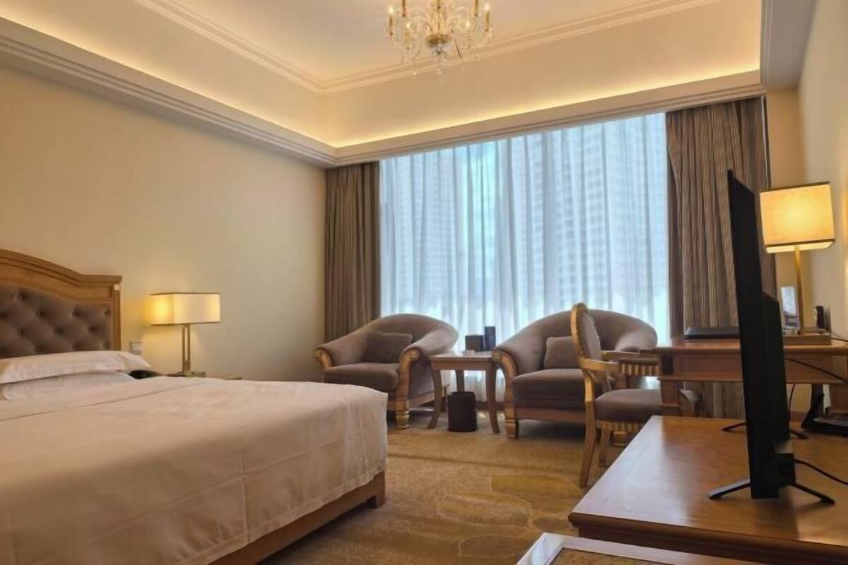 A hotel room with a large bed, two armchairs, a desk with a computer, and a TV.