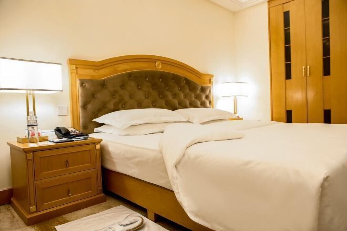 Una habitación de hotel con una cama grande, cabecero de madera, mesitas de noche con lámparas y un armario.