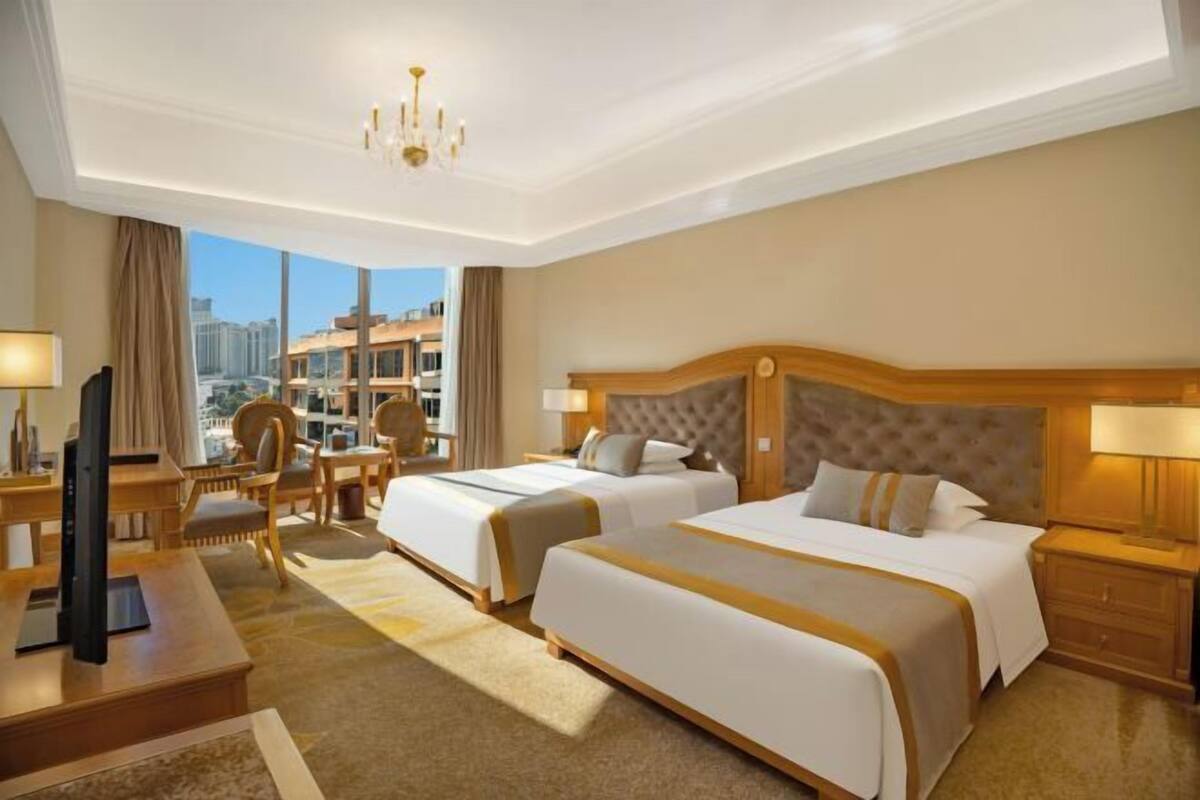 A hotel room with two beds, a desk, a chair, a TV, and a view of the city.