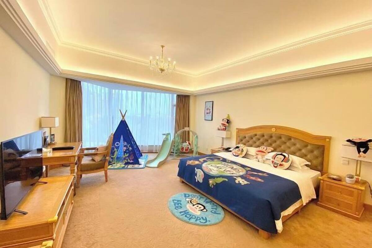 A hotel room with a bed, a desk with a TV, a chair, a small play area with a slide, and a window with curtains.