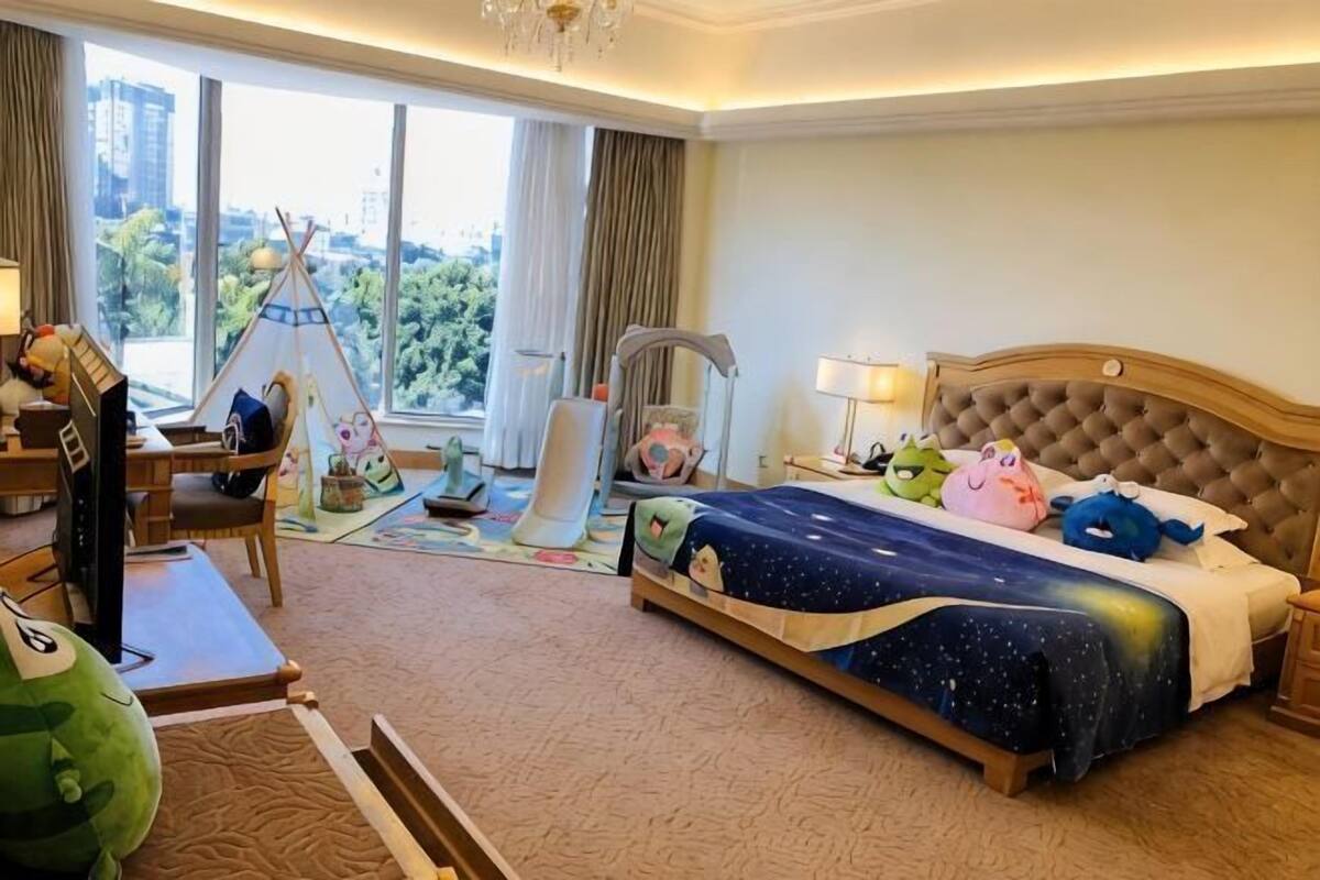 A hotel room with a bed, a desk, a chair, a window with a view, and a tent.