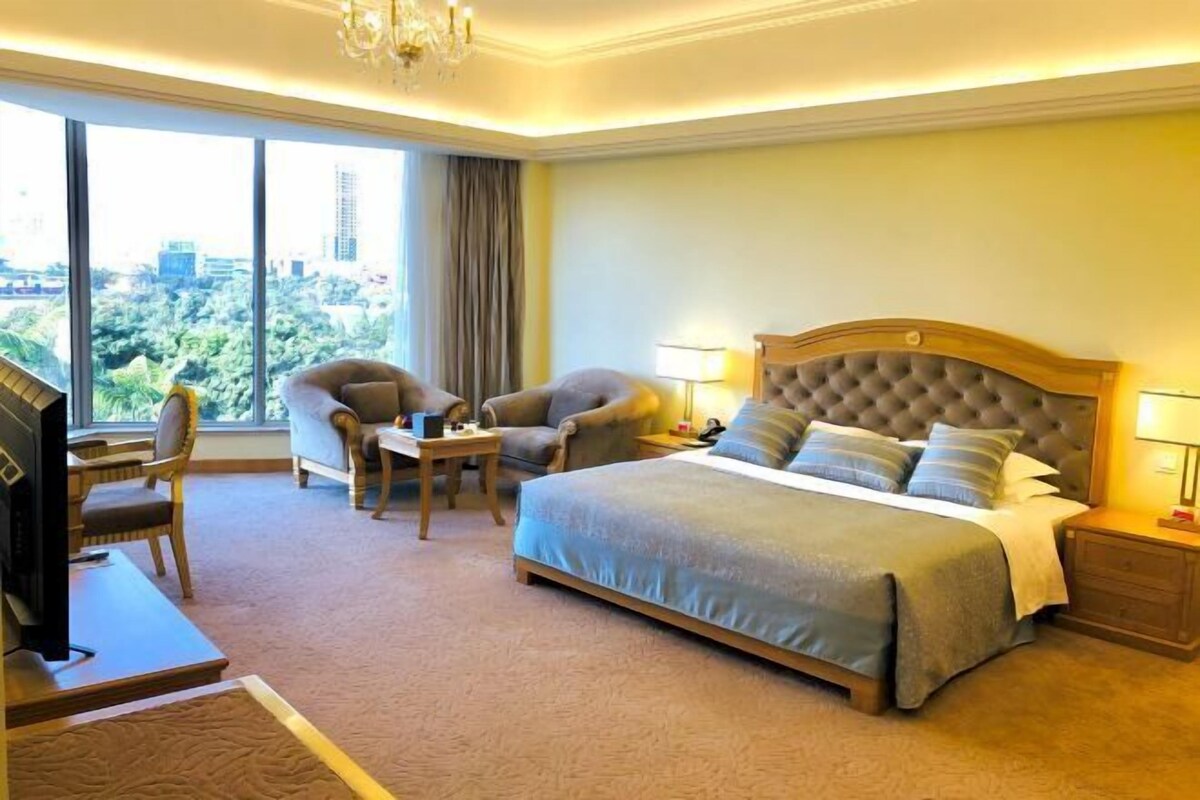 Habitación de hotel con una cama grande, dos sillones, un escritorio con televisión y vistas a la ciudad a través de amplios ventanales.