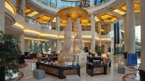 Um hall amplo com uma escultura decorativa central, diversas áreas de estar e um teto alto com claraboias.