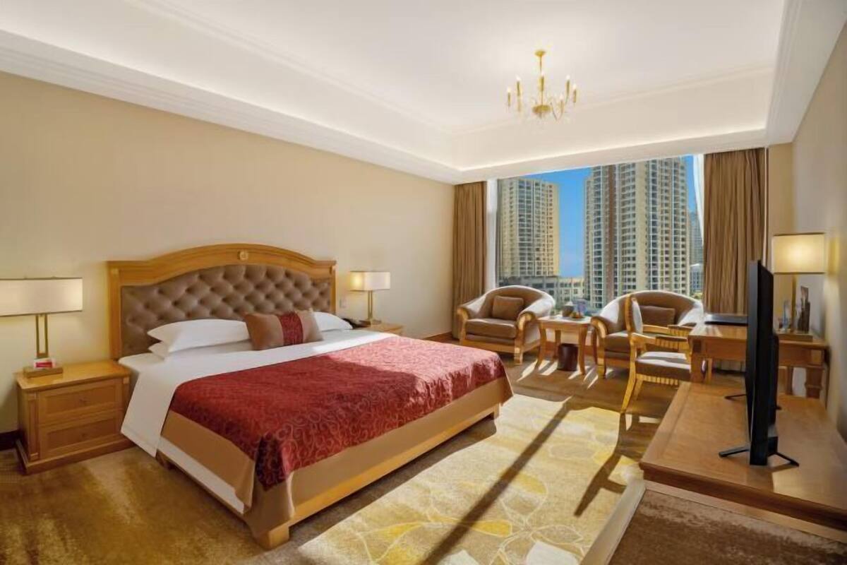 A hotel room with a large bed, a television, a desk, and a view of tall buildings.