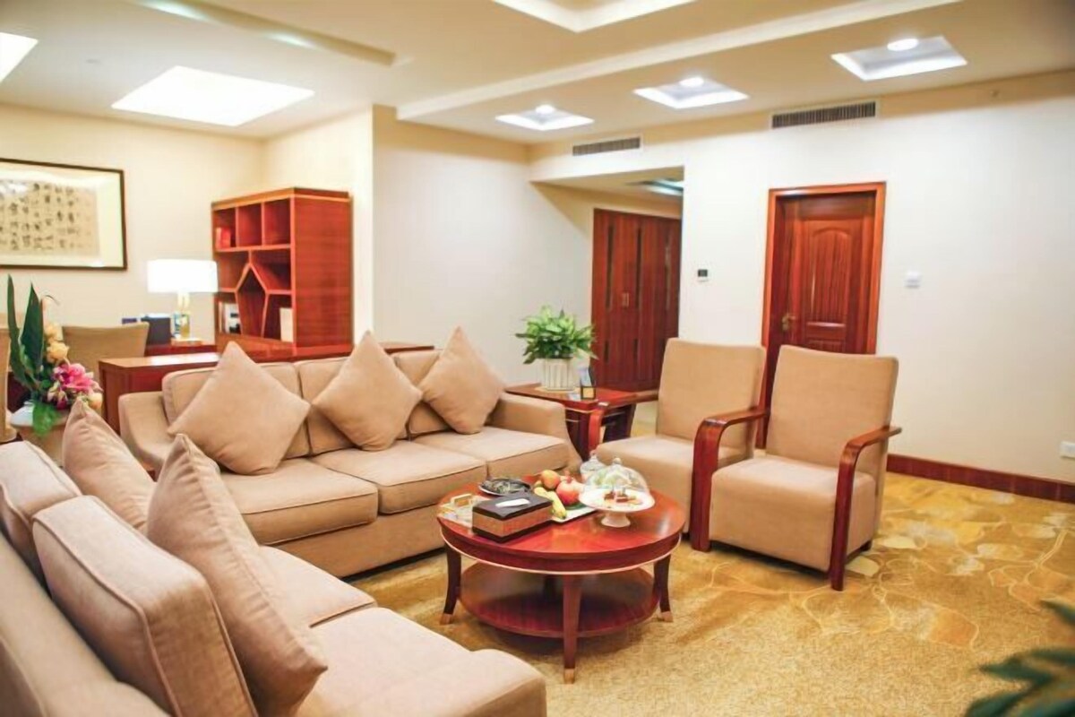 Una sala de estar con un sofá color beige, una mesa de centro redonda y un sillón de madera. Se observa una estantería y una puerta al fondo.