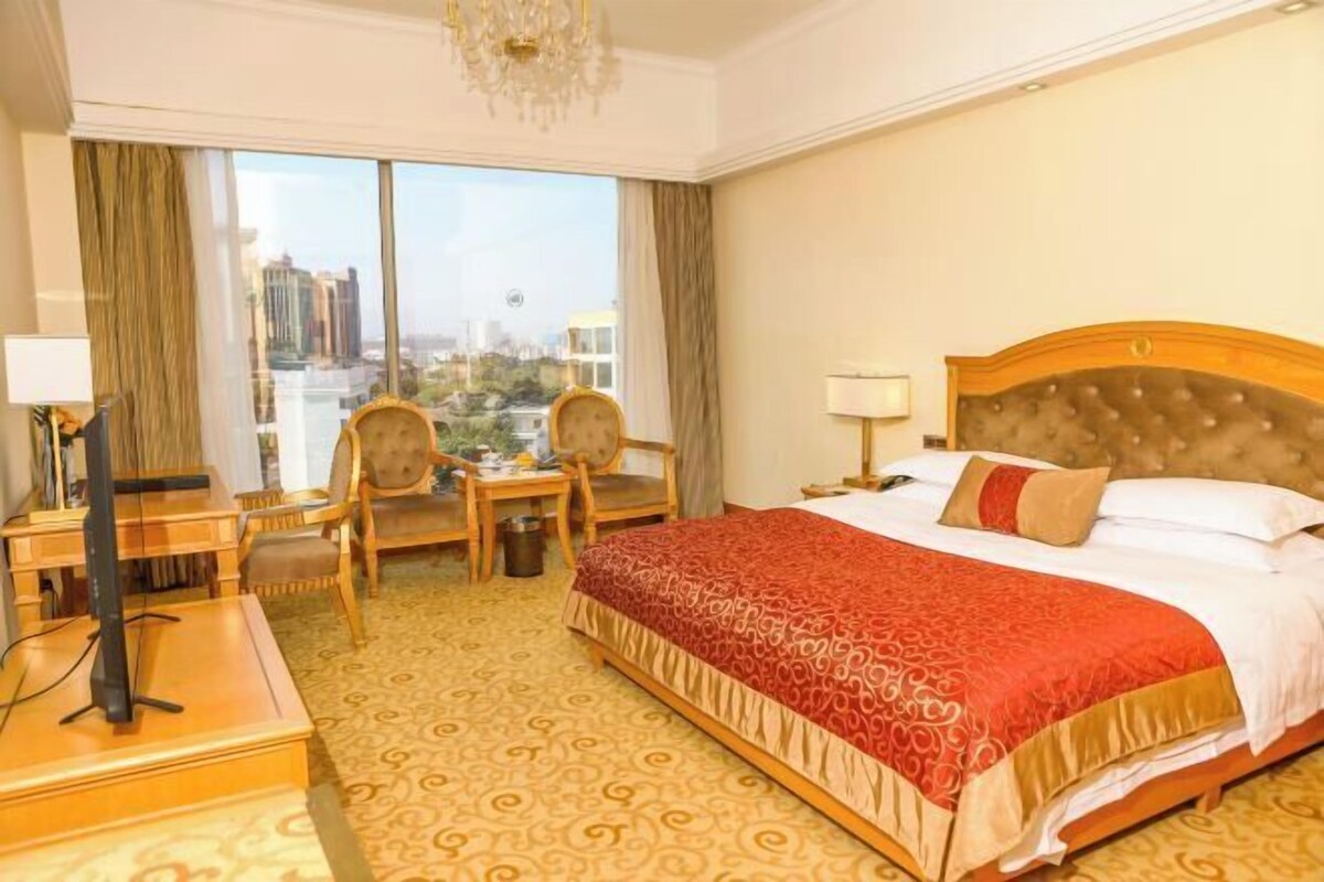 Habitación de hotel con una cama grande, un escritorio con silla, un televisor y vistas a la ciudad a través de amplios ventanales.