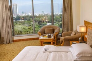 A hotel room with a large window, a bed, two armchairs, a small table with a tray, and a lamp.